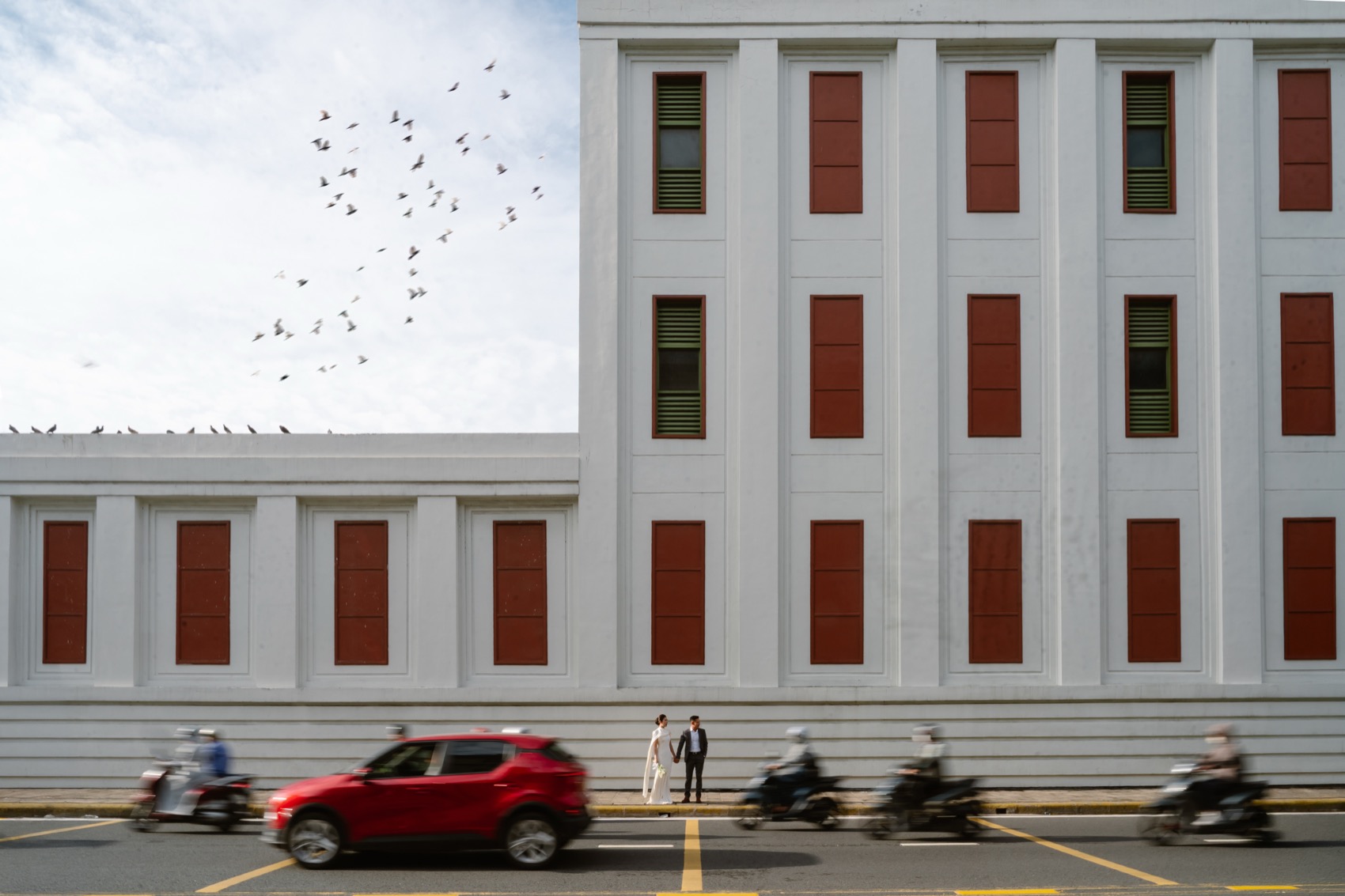 Couple by a red-shuttered heritage building as birds take flight in Saigon