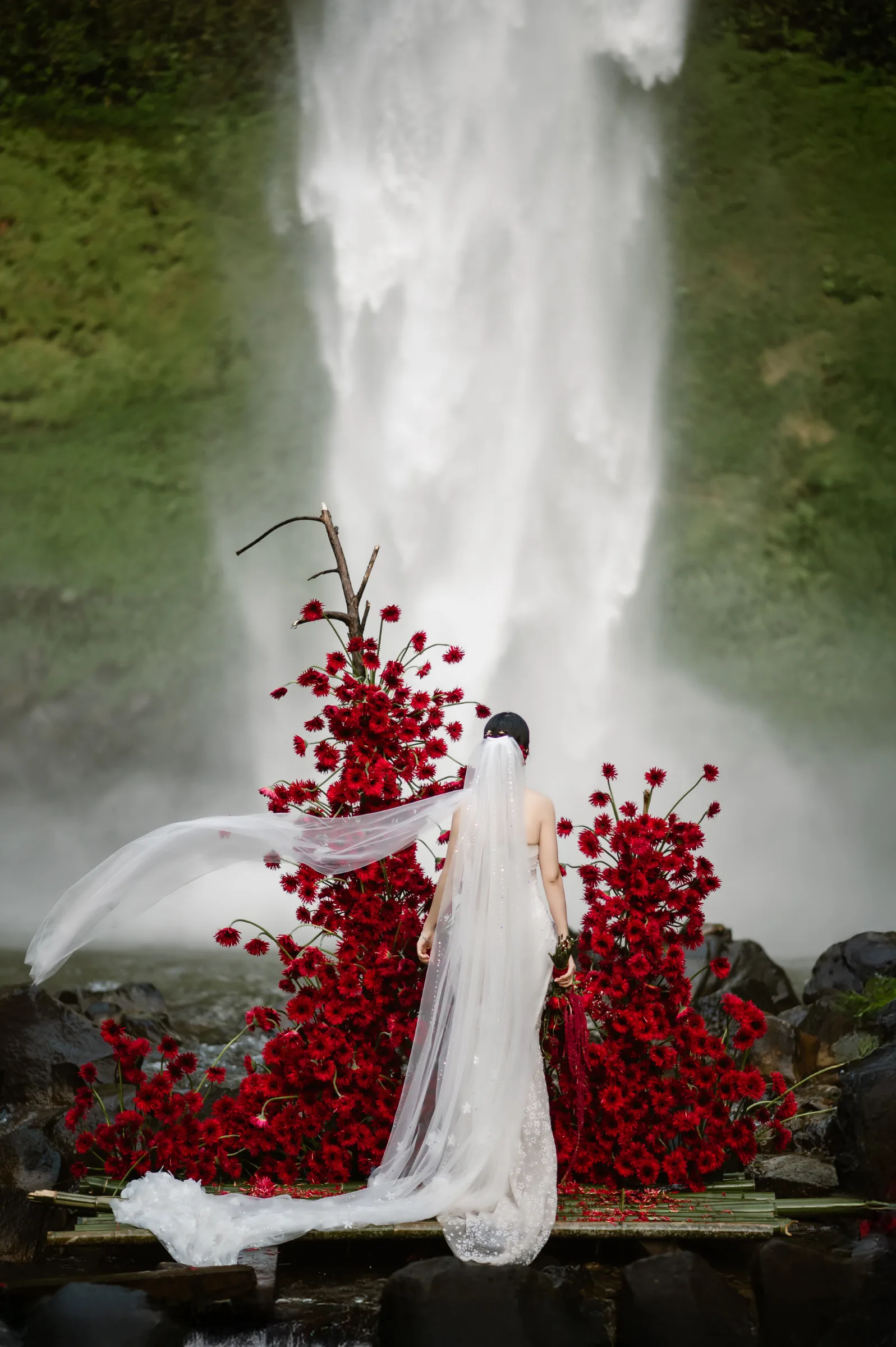 Bride with a red-flower train at a waterfall during an adventure elopement in Vietnam