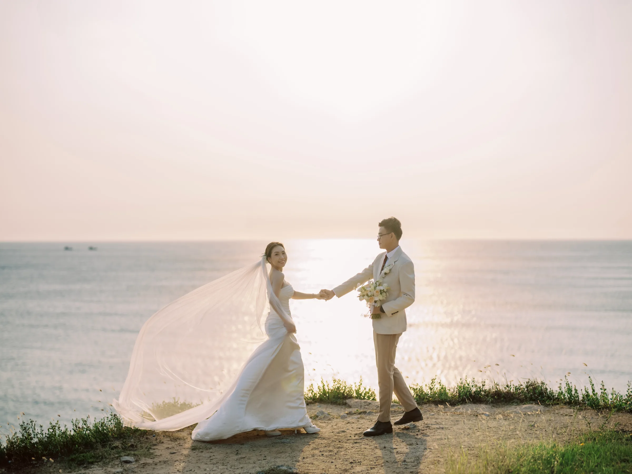 Couple holding hands on a clifftop as the bride's veil flies, Phu Yen pre-wedding
